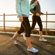 Two people in athletic wear power walk on a paved ocean path, sunlight behind them. Only their legs and torsos are visible—showing a smart way to build running distance and avoid burnout while enjoying scenic exercise. Austin Marathon Half Marathon & 5K