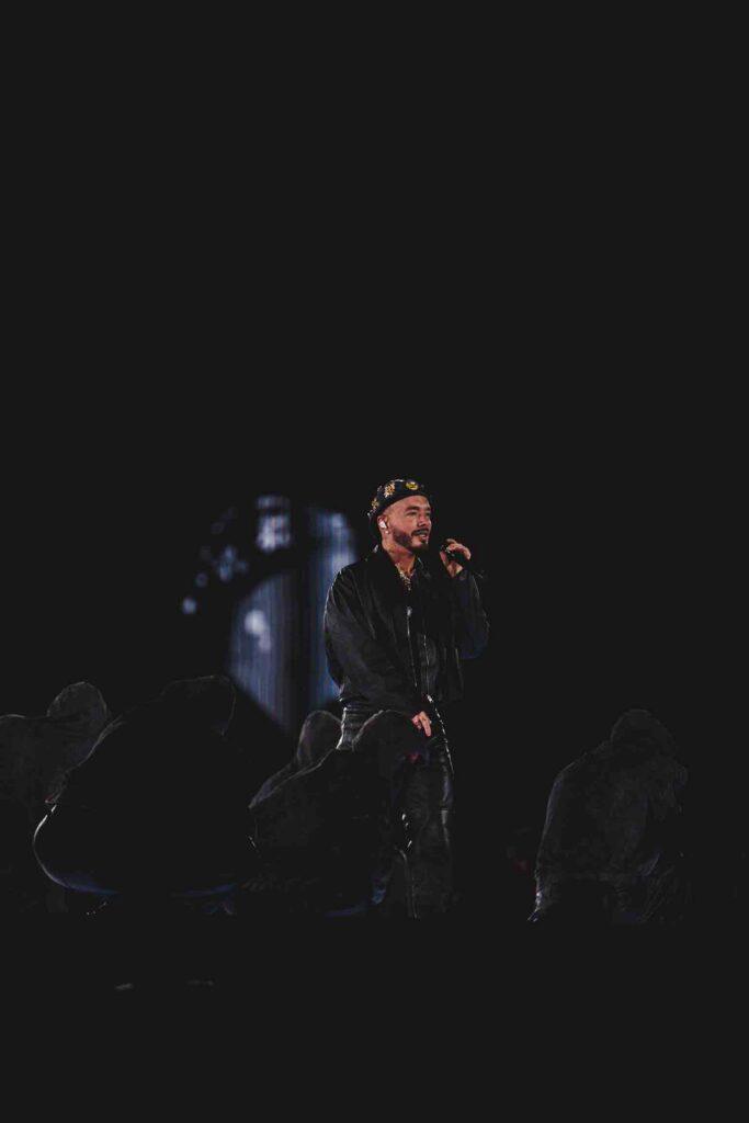 Male performer on a dark concert stage, microphone in hand, audience silhouettes at the foreground.