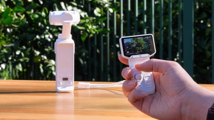 The Insta360 Luna Ultra set down in the background, with a hand holding its detached remotein the foreground.
