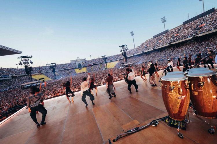 Wide view of a packed outdoor stadium concert with dancers on stage and a sea of cheering fans in the stands; foreground shows large drums onstage.