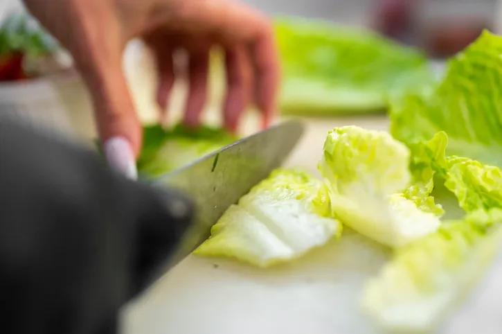 A person uses a chef's knife to chop fresh, bright green romaine lettuce leaves on a white cutting board, preparing ingredients in a bright kitchen.