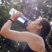 A woman in athletic wear drinks water from a blue bottle outdoors, sunlight filtering through trees behind her. With a braid and fitness tracker, she embodies summer fitness on a refreshing outdoor workout. Austin Marathon Half Marathon & 5K