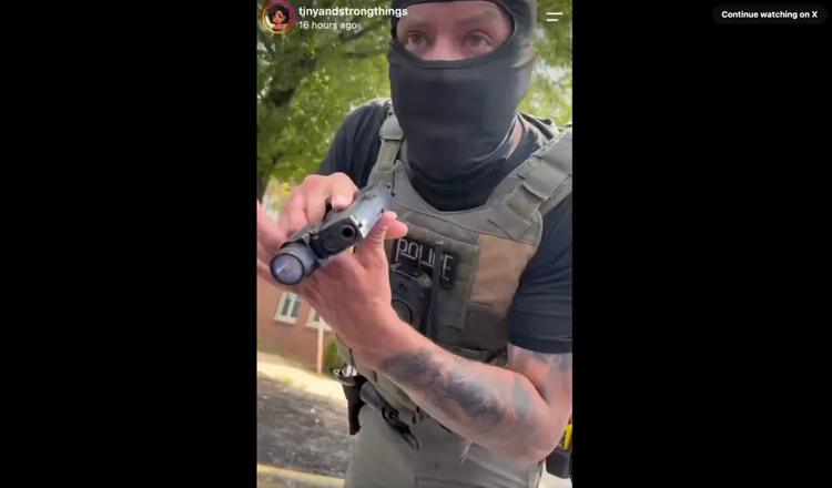 A person wearing a black mask and tactical gear, holding a police-style firearm.