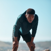 A young man in athletic clothing leans forward with his hands on his knees, appearing tired or catching his breath outdoors—pushing through the Summer Consistency Challenge to stay on track beneath a clear sky and blurred landscape. Austin Marathon Half Marathon & 5K
