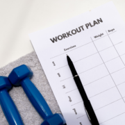 A workout plan sheet with columns for exercises, weight, and reps lies on a gray towel next to two blue dumbbells and a black pen—perfect for those looking to build running distance while tracking progress throughout the week. Austin Marathon Half Marathon & 5K