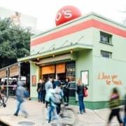People walk and cycle past a green building with a red “O’s” sign and “i love you so much.” painted on the side—a favorite for post-run meetups and one of the best coffee shops in Austin, with outdoor seating and a welcoming vibe. Austin Marathon Half Marathon & 5K