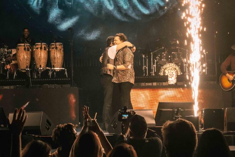 Two performers embrace on a concert stage as fireworks shoot overhead, with a drummer on the left and cheering audience in front.