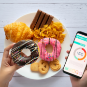 A person holds a plate with a croissant, chocolate, donuts, cookies, and cornflakes in one hand, and a smartphone displaying a calorie counter app reading 1200 calories in the other—refuel recharge after your Austin recovery day. Austin Marathon Half Marathon & 5K