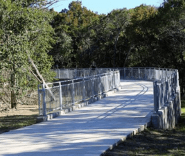 A paved, elevated walkway with metal railings on both sides curves gently to the left at Walnut Creek Park in Austin Texas. The path is surrounded by dense trees with green foliage, highlighting Austin’s hidden running routes. Shadows from the trees fall across the walkway on a sunny day. Austin Marathon Half Marathon & 5K
