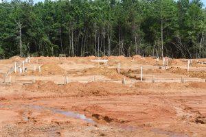 A cleared construction site with red dirt, wooden forms, white pipes and small mounds of soil in front of a tree line.