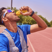 A man in sports gear and sunglasses drinks from a yellow pouch on a running track, focusing on marathon fueling amid trees and clear sky in the background. Austin Marathon Half Marathon & 5K