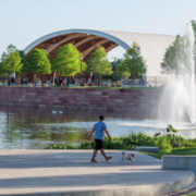 A person enjoys an Austin morning exercise, walking a small dog by a pond with a fountain in a park. In the background, people gather under a large, curved pavilion surrounded by trees. Austin Marathon Half Marathon & 5K
