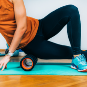 A person wearing an orange tank top, black leggings, and blue sneakers uses a foam roller on their thigh while sitting on a turquoise exercise mat indoors to refuel after a run, perfect for Austin Recovery Day. Austin Marathon Half Marathon & 5K