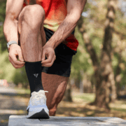 A person in athletic wear kneels outdoors, adjusting their black sock—a vital part of their running gear. The background is blurred, showing trees and sunlight. The person wears a watch and white running shoes, appearing ready to tick another item off their official training checklist. Austin Marathon Half Marathon & 5K