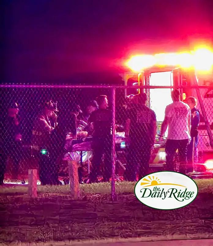 Nighttime scene with a crowd behind a chain-link fence, illuminated by bright pink/red lights from a vehicle on the right; a 'The Daily Ridge' logo is visible in the bottom corner.
