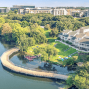 A boardwalk curves along a lakeshore near grassy lawns, trees, and townhouses—an inviting route for East Austin running enthusiasts or members of local running clubs. Colorful kayaks and paddleboards are visible on the grass, with apartment buildings in the background under a clear blue sky. Austin Marathon Half Marathon & 5K