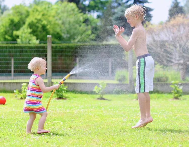 Kid and toddler playing with a garden hose in the backyard on a hot summer day, enjoying outdoor water play and cooling off together.