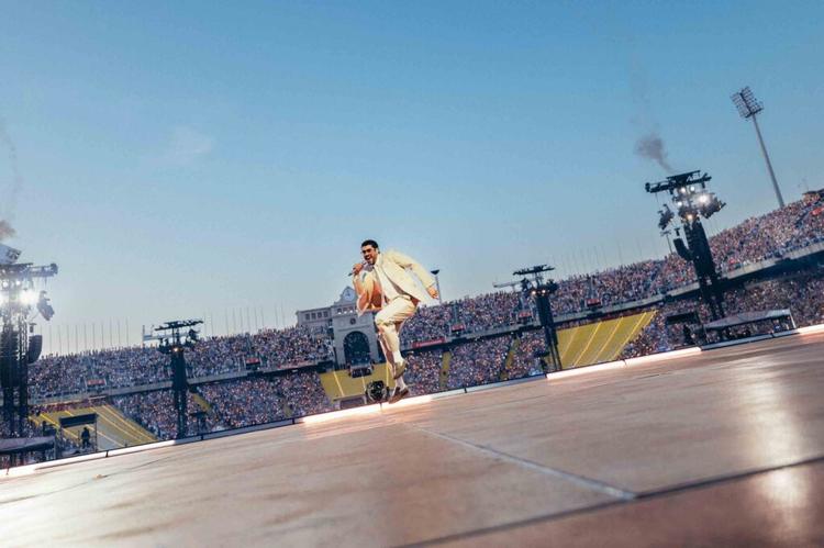 Male singer in a beige suit performs mid-air on a stadium stage, with a large crowd watching.