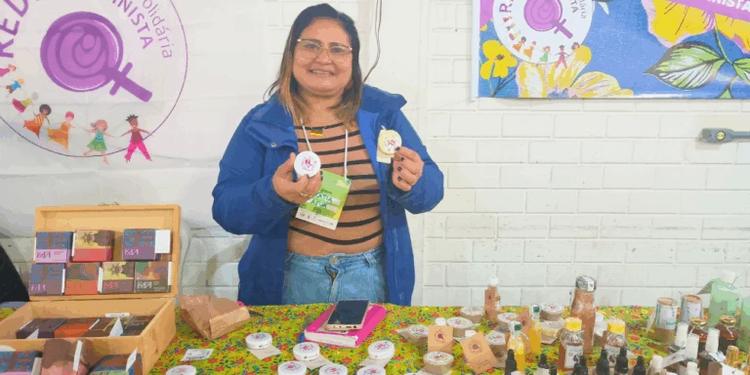 Foto colorida de Katia Pantoja. Ela é uma mulher branca, com cabelo preto com mechas loiras, liso. Ela sorri e segura potes de pomadas.