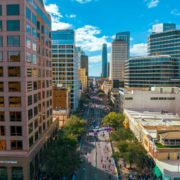 Aerial view of a city street surrounded by tall modern buildings, with a large crowd gathered below for a mid-year fitness check on a sunny day. The sky is blue with a few clouds, and trees line the sidewalks. Austin Marathon Half Marathon & 5K