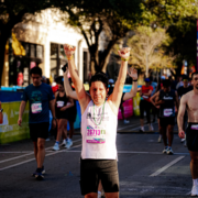 A marathon runner wearing a white tank top and bib number 26713 raises both arms in triumph at the finish line, celebrating a February goal and inspiring others on their mid-year fitness check, surrounded by fellow runners on a sunlit city street. Austin Marathon Half Marathon & 5K