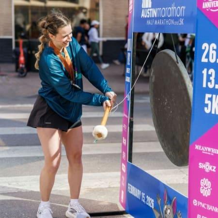 A woman smiles as she strikes a large gong with a mallet at the Austin Marathon and 5K finish area. Wearing a medal, teal jacket, and black shorts, she celebrates what's next. Event banners and people fill the background. Austin Marathon Half Marathon & 5K
