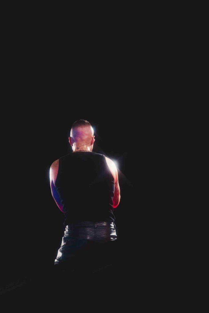 Back view of a muscular man in a black sleeveless shirt on a dark stage with colorful lighting behind him