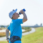 A man in a blue shirt and cap drinks water from a bottle while standing outdoors on a grassy path under a clear sky, taking a break during his running training to avoid burnout. He is wearing a waist pouch and sunglasses on his cap. Austin Marathon Half Marathon & 5K