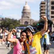 Two running crew members celebrate with medals and raised fists in front of the Texas State Capitol building. Austin Marathon Half Marathon & 5K