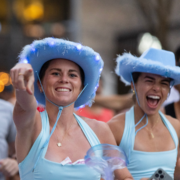 Two women wearing light blue cowboy hats and matching outfits are smiling and celebrating at an outdoor event. One points at the camera as they cheer excitedly during the 24 Weeks countdown to the Austin Marathon, their hats glowing with lights and feather trim. Austin Marathon Half Marathon & 5K