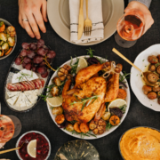 A festive table set with roasted chicken, vegetables, cheese, grapes, and charcuterie awaits—perfect for an Austin Recovery Day or to Run Refuel Recharge with friends. Two hands toast with wine amid cranberries, mushrooms, and drinks. Austin Marathon Half Marathon & 5K