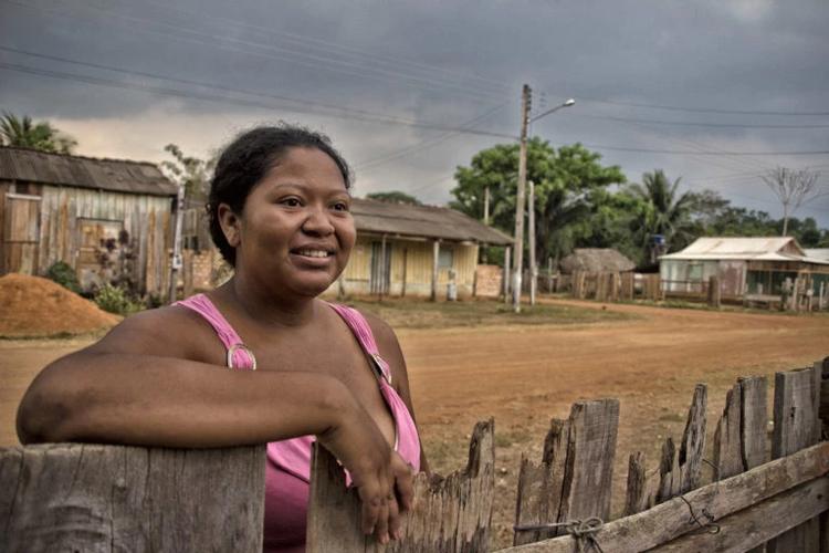 Foto colorida de mulher negra escorada em cerca de madeira. Ela sorri e olha para frente. Ao fundo, casas e postes de energia. Comunidade Quilombolas do Forte Príncipe da Beira, no Vale do Guaporé, em Costa Marques, em Rondônia. (Foto: Marcela Bonfim/Amazônia Real/2018).