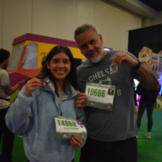 A smiling young woman and an older man proudly display their race medals and bibs after an exciting Race Weekend, standing indoors with colorful decor and a van in the background. The woman’s bib reads “Cheyenne 14868” and the man’s “19686.”. Austin Marathon Half Marathon & 5K