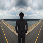 A man in a suit stands at a forked road, facing a large water reservoir under a cloudy sky, symbolizing a decision point—much like reaching your February goal or reflecting during a Mid-Year Fitness Check. Austin Marathon Half Marathon & 5K