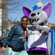 A smiling marathon finisher poses with a large bat mascot in an 