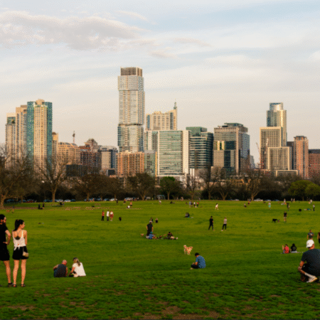People unwind and walk dogs on a large grassy park with scattered trees, making it ideal for a day trip, while a modern city skyline with tall buildings rises in the background under a partly cloudy sky. Austin Marathon Half Marathon & 5K