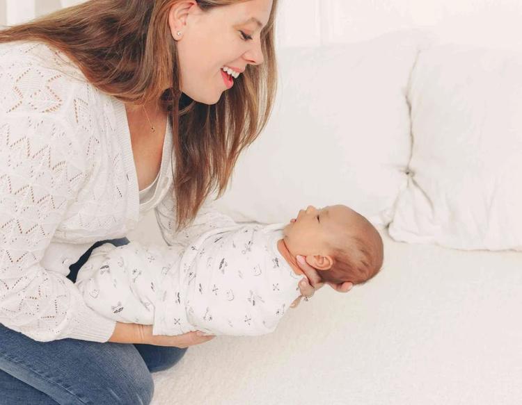 Mom gently laying her newborn on the bed wrapped in a KeaBabies Lyra Muslin Swaddle Blanket.