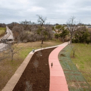 A person jogs on a winding red path through a park in East Austin, while another person walks nearby. Leafless trees surround the area, and a railroad track runs parallel to the path under a cloudy sky—a familiar scene for local running clubs before heading out for post-run tacos. Austin Marathon Half Marathon & 5K