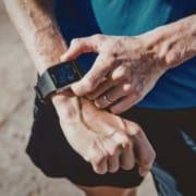 Close-up of a person in athletic clothing adjusting a black sports smartwatch on their wrist, with the screen displaying fitness tracking info—a perfect moment for any summer running guide. The blurred background highlights focus and determination. Austin Marathon Half Marathon & 5K
