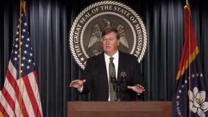 Gov. Tate Reeves stands at a podium between two flags and gestures with one hand. Behind him is the Great Seal of the State of Mississippi.