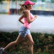 A woman wearing a pink tank top, pink cap, and gray shorts jogs outdoors on a paved path lined with greenery, embodying the spirit of a Summer Running guide as she smiles and stays active in the sunshine. Austin Marathon Half Marathon & 5K