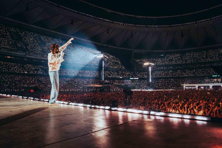 Performer on a large stadium stage raises an arm toward the cheering crowd, thousands of tiny lights filling the audience.