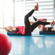 Two people exercise on foam rollers in a bright fitness studio, incorporating gym workouts into their routines for enhanced longevity. One lies on her back with arms extended and legs raised, wearing a red tank top and black leggings. A large red exercise ball is in the foreground. Austin Marathon Half Marathon & 5K