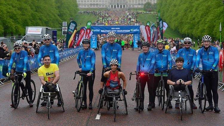 The picture shows three people on wheelchairs in front of other people dressed in blue on bicycles. Stormont and hundreds of marathon runners are behind them  