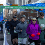 Runners in athletic gear participate in the Austin Marathon & Half Marathon, passing under a blue and green start banner. A woman in a bright pink 