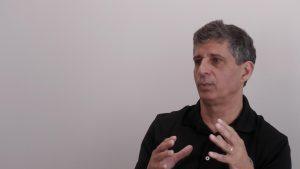 A short-haired man wearing a black polo shirt gestures while speaking to someone outside the frame.