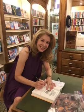 Author Melissa Roske smiling and signing a book at a bookstore event. She is seated at a wooden table in front of bookshelves, wearing a sleeveless purple dress.