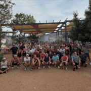 Large group of people posing together outdoors on dirt ground, many wearing athletic clothes, hats, and sunglasses—typical of East Austin running clubs. Trees and a shaded structure are visible in the background under a cloudy sky, setting the perfect scene for post-run tacos. Austin Marathon Half Marathon & 5K