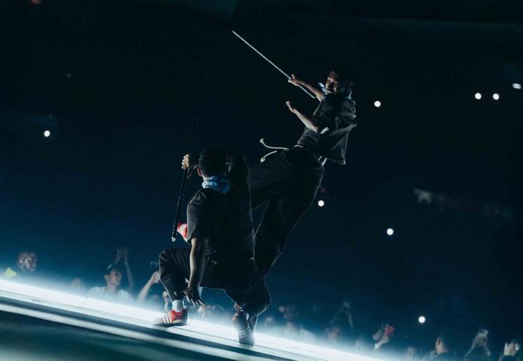 Two performers balance on a glass stage in a dark arena, holding long poles as bright lights twinkle in the background.]