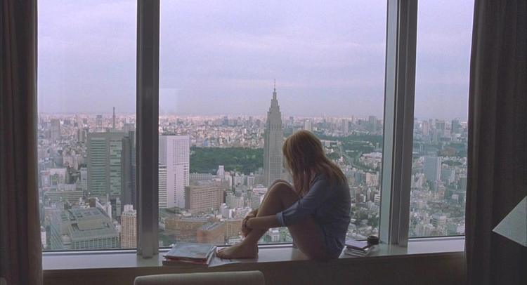 A woman sitting in front of the window, gazing at the panoramic view of the city and the clock tower in the background at dusk.